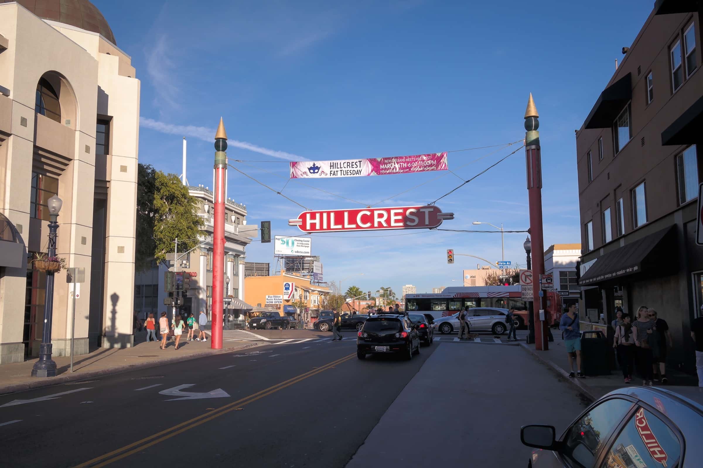 The red and white Hillcrest sign hanging between two red poles over University Avenue, with storefronts, a city bus and people on the sidewalks