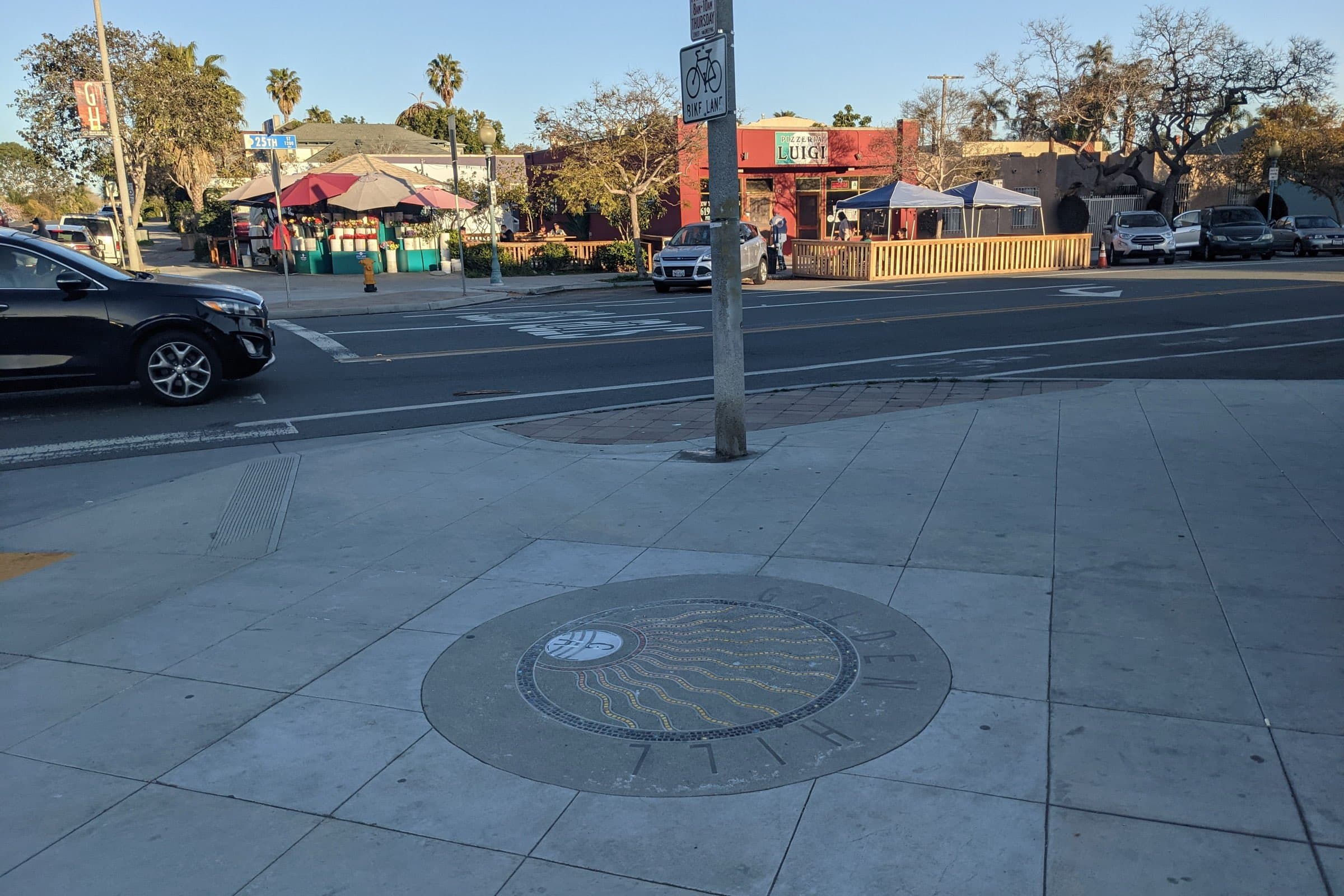 A Golden Hill mosaic medallion in the sidewalk at 25th Street, across from a corner flower stand, a restaurant patio and palm trees in late-afternoon sun